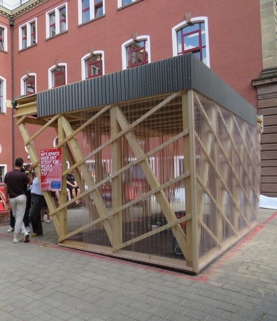 Außenansicht eines Pavillons aus Holz, durchsichter Plexiglas-Fassade und Wellblechattika mit umstehenden Personen