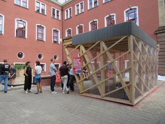 Außenansicht eines Pavillons aus Holz, durchsichter Plexiglas-Fassade und Wellblechattika mit umstehenden Personen