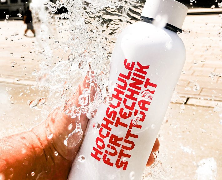 Das Bild zeigt eine weiße 05, Liter Trinkflasche mit einem roten Aufdruck "Hochschule für Technik Stuttgart". Wassertropfen spritzen um die Flasche herum.
