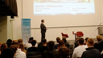 Ein Redner auf der Bühne der Aula zeigt eine Präsentation