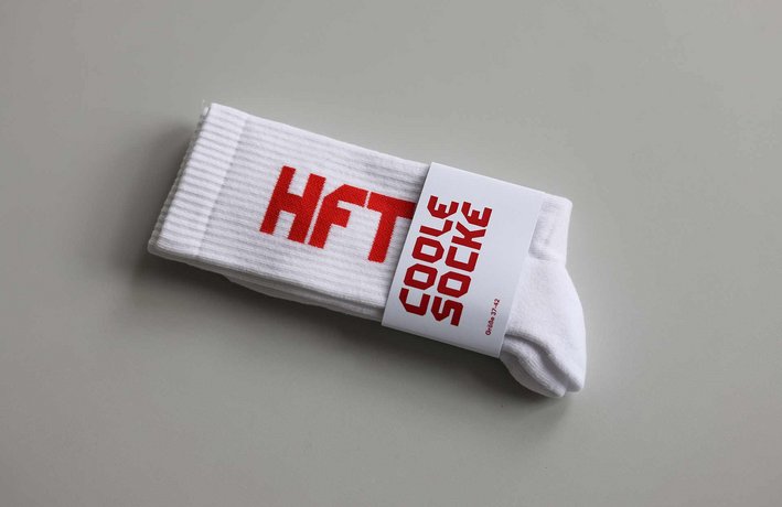Tennissocke mit Stick "HFT". Als Verpackung ist eine Banderole aus Pappe um die Socken gelegt mit einem Aufdruck "Coole Socke".