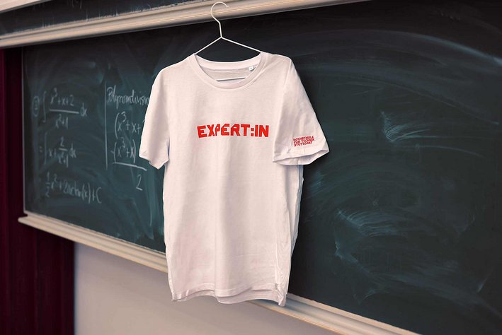Weißes Tshirt mit der Aufschrift "Expert:in" in HFT Stuttgart Schrift. Rotes Logo auf linkem Ärmel. Das Tshirt hängt auf einem Kleiderbügel an einer Tafel in einem Vorlesungsraum.