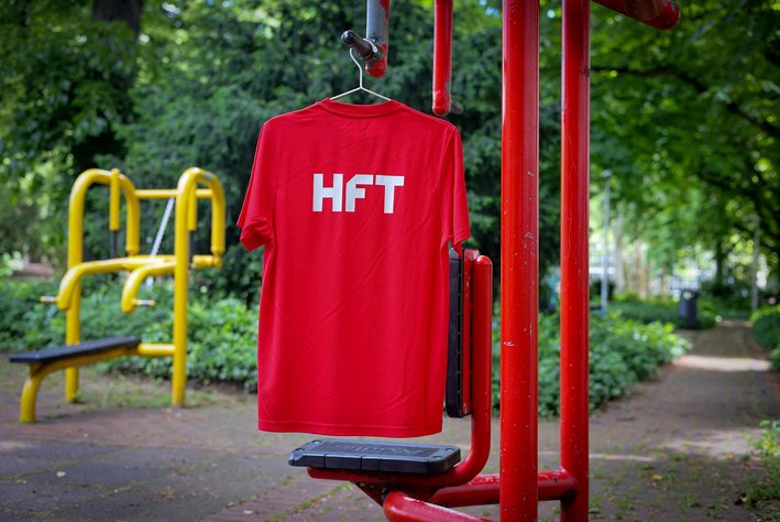 Ein rotes HFT Sportshirt mit weißem Aufdruck "HFT" hängt an einem Fitnessgerät im Park