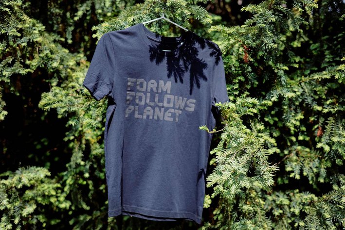 Ein schwarzes Tshirt mit schwarzem Druck "Form follows Planet" ist auf einem Kleiderbügel an einem Busch aufgehängt