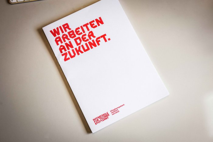 Weißer DIN A4 Block mit der Aufschrift "Wir arbeiten an der Zukunft" und dem HFT Stuttgart Logo in rot