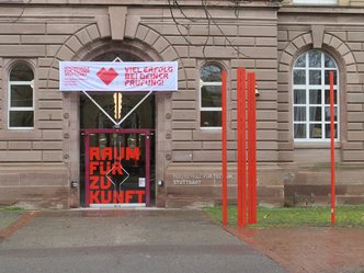 Eingang des Hauptgebäudes mit Beklebung "Raum für Zukunft", rechts daneben vier rote Stahlstelen.